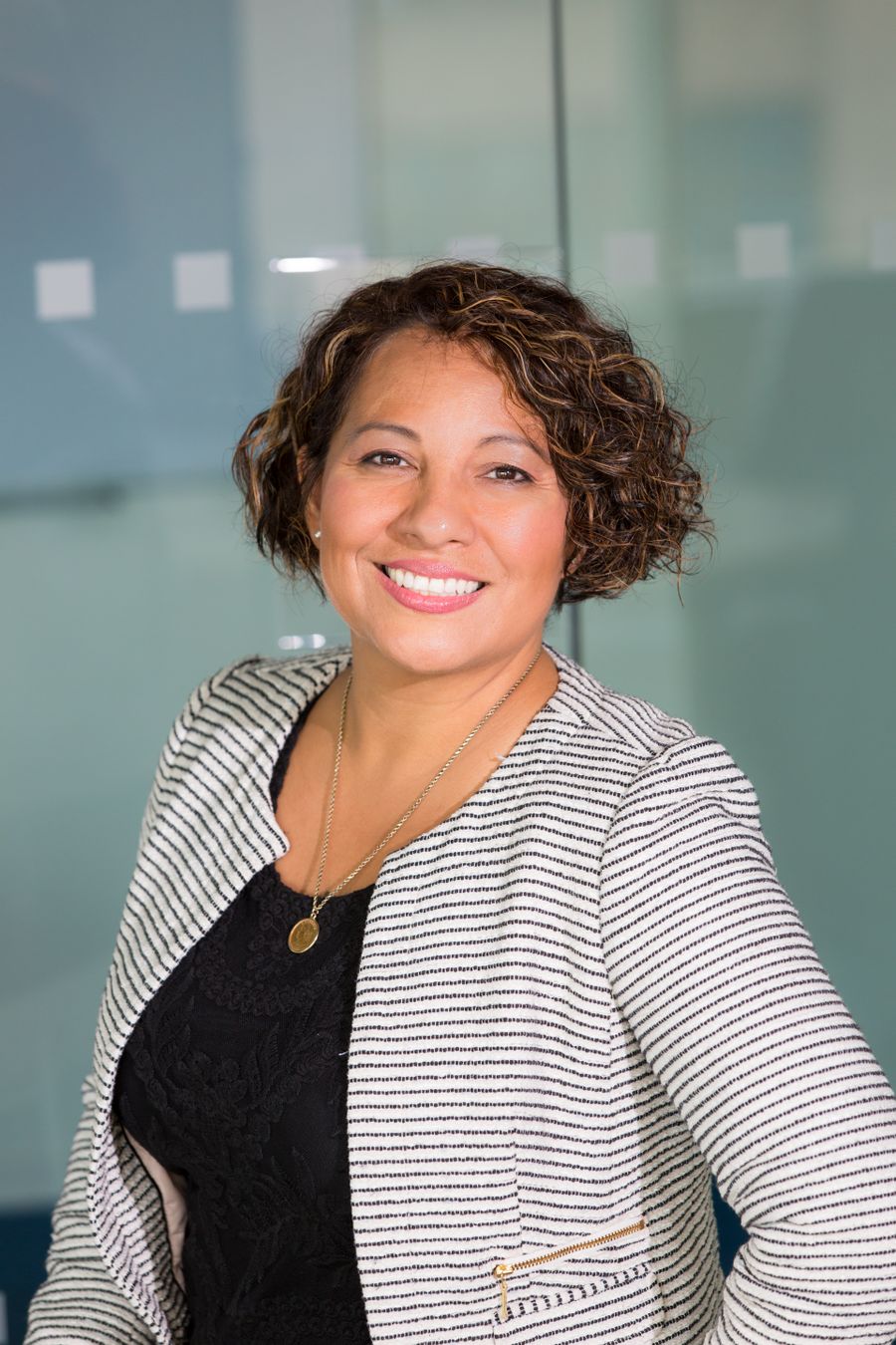 A smiling woman in a striped jacket, standing in a glass-walled office